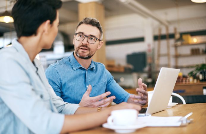 two men talking over a laptop and cup of coffee